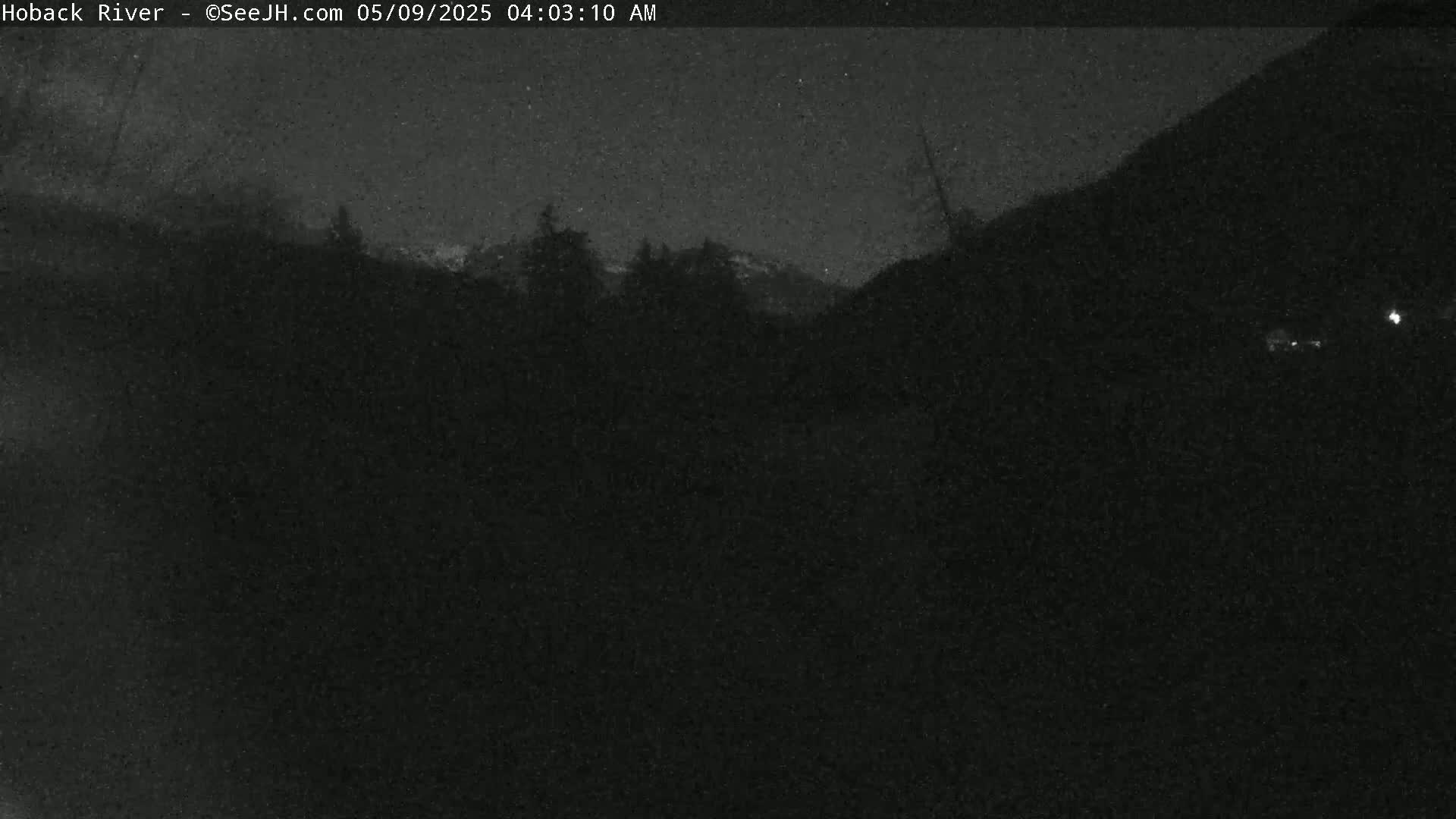 A nighttime, low-light outdoor image shows a dark landscape with a silhouetted line of trees against a darker mountain range.