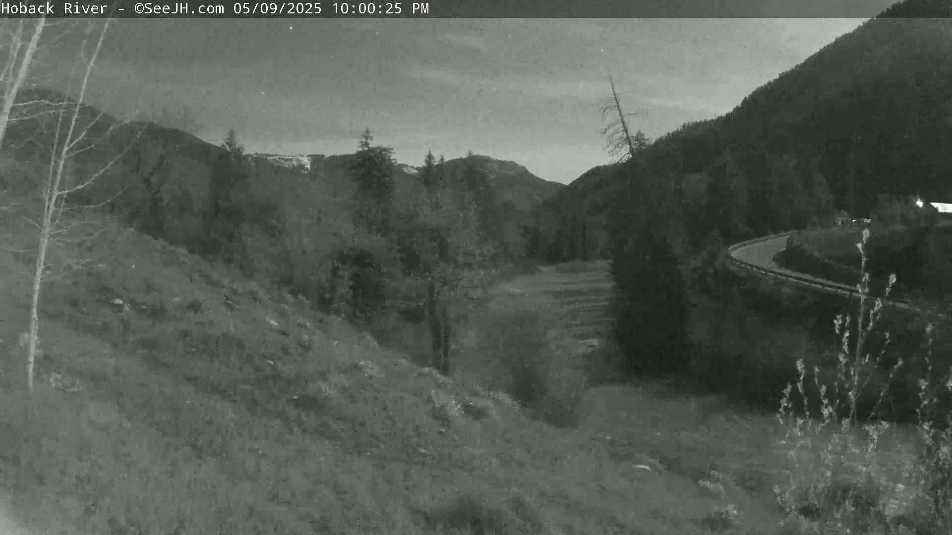A nighttime, grayscale view of a winding road curving through a mountainous valley under a dusky sky.