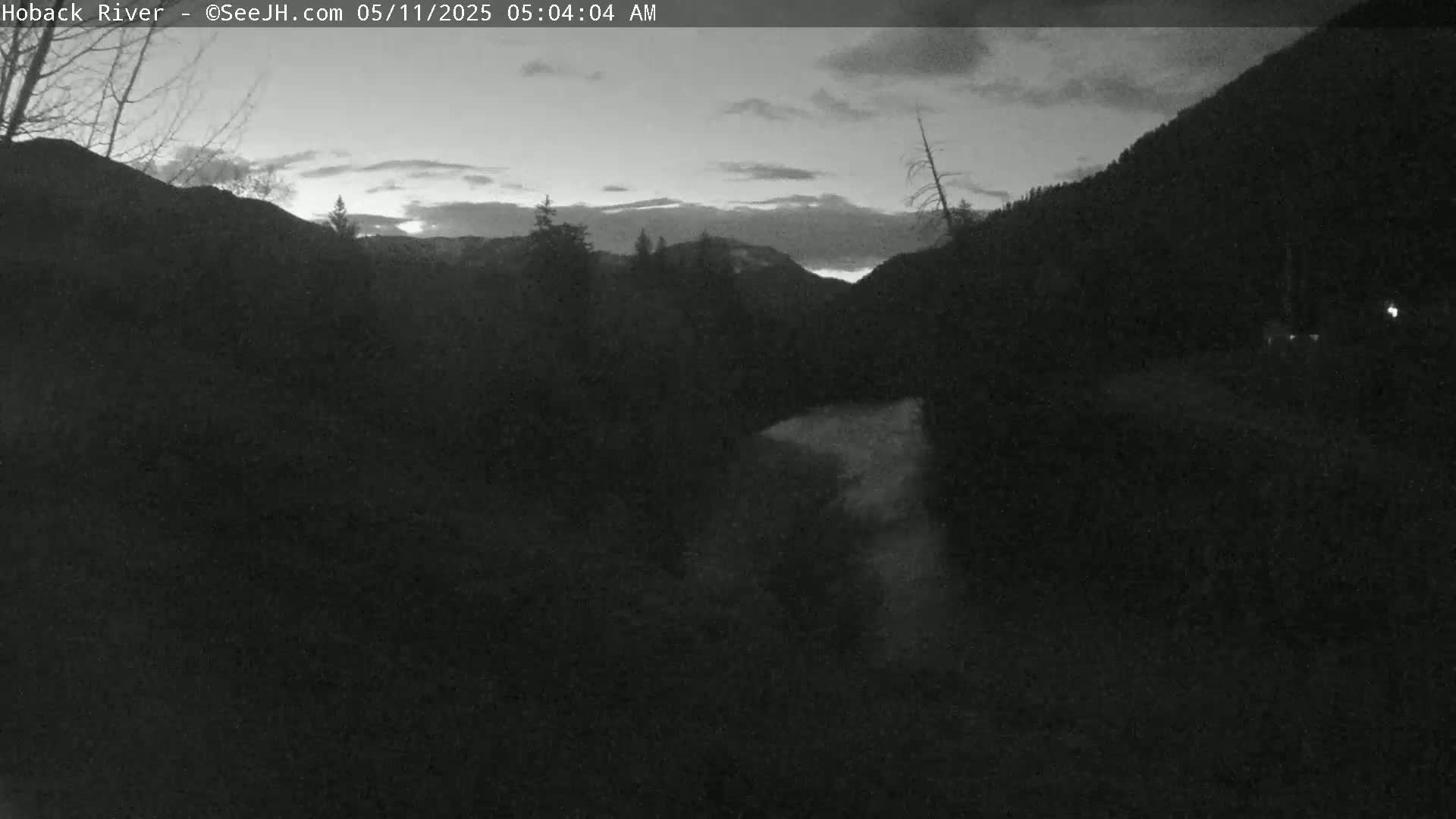 A dark, pre-dawn scene shows a river valley nestled between mountains under a cloudy sky.