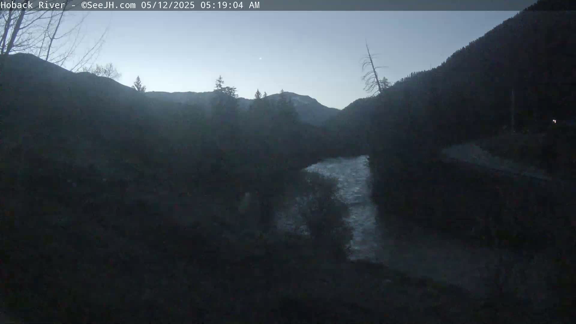 A dark, pre-dawn view of a river flowing through a valley, framed by mountains and trees under a clear, twilight sky.