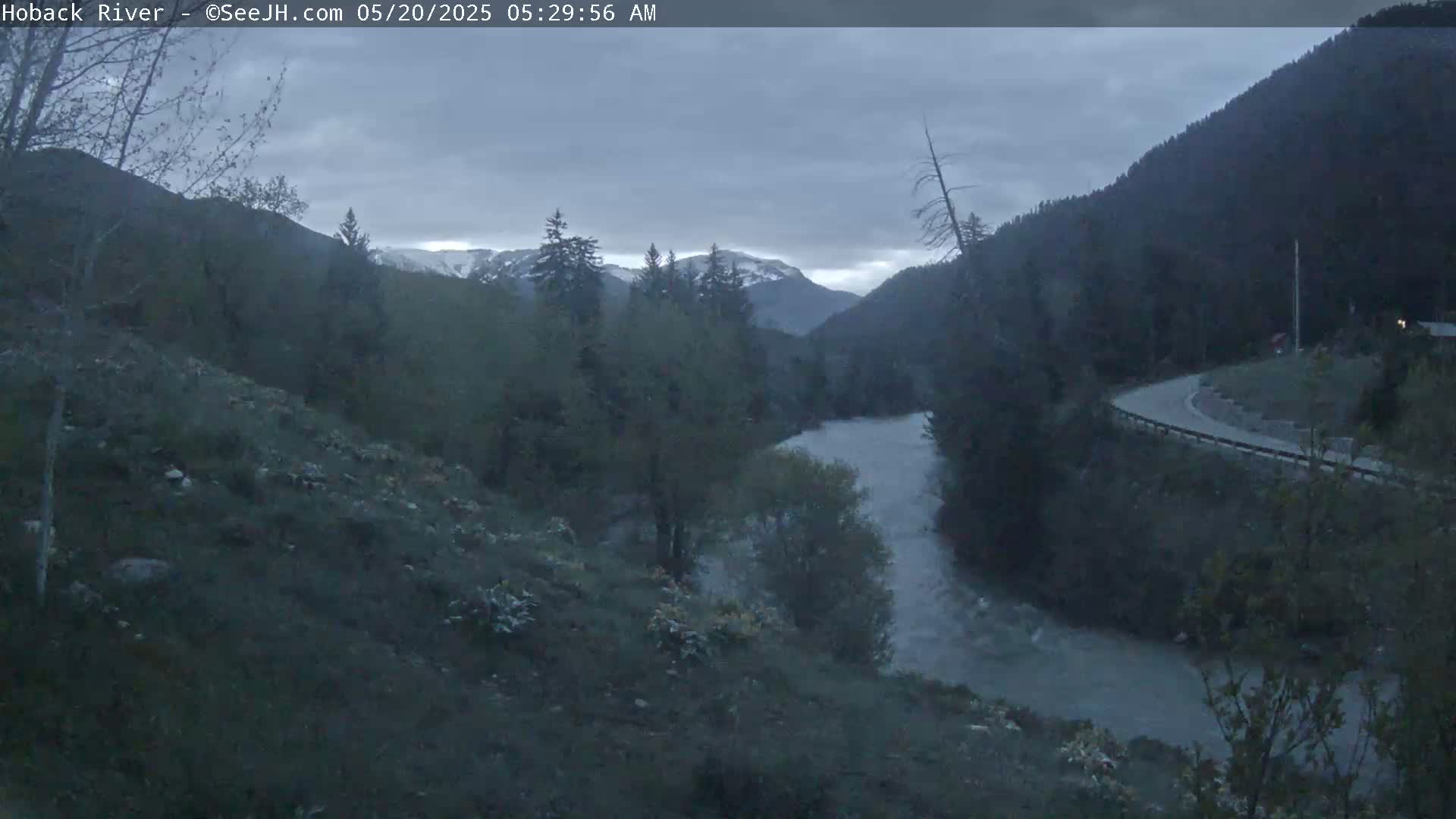 A river flows through a valley, under an overcast sky, between mountains with some snow cover.