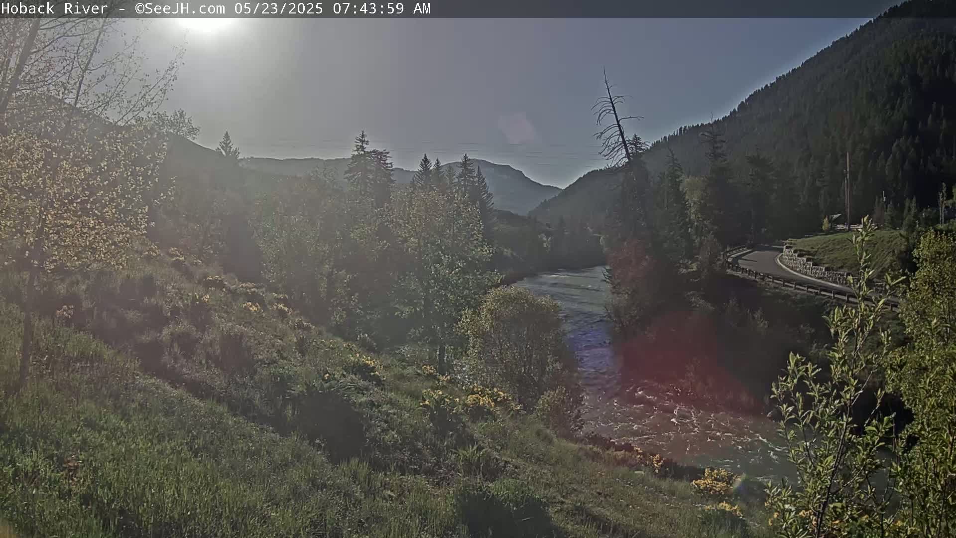 A river flows through a valley under a mostly clear sky, surrounded by green hillsides and trees.