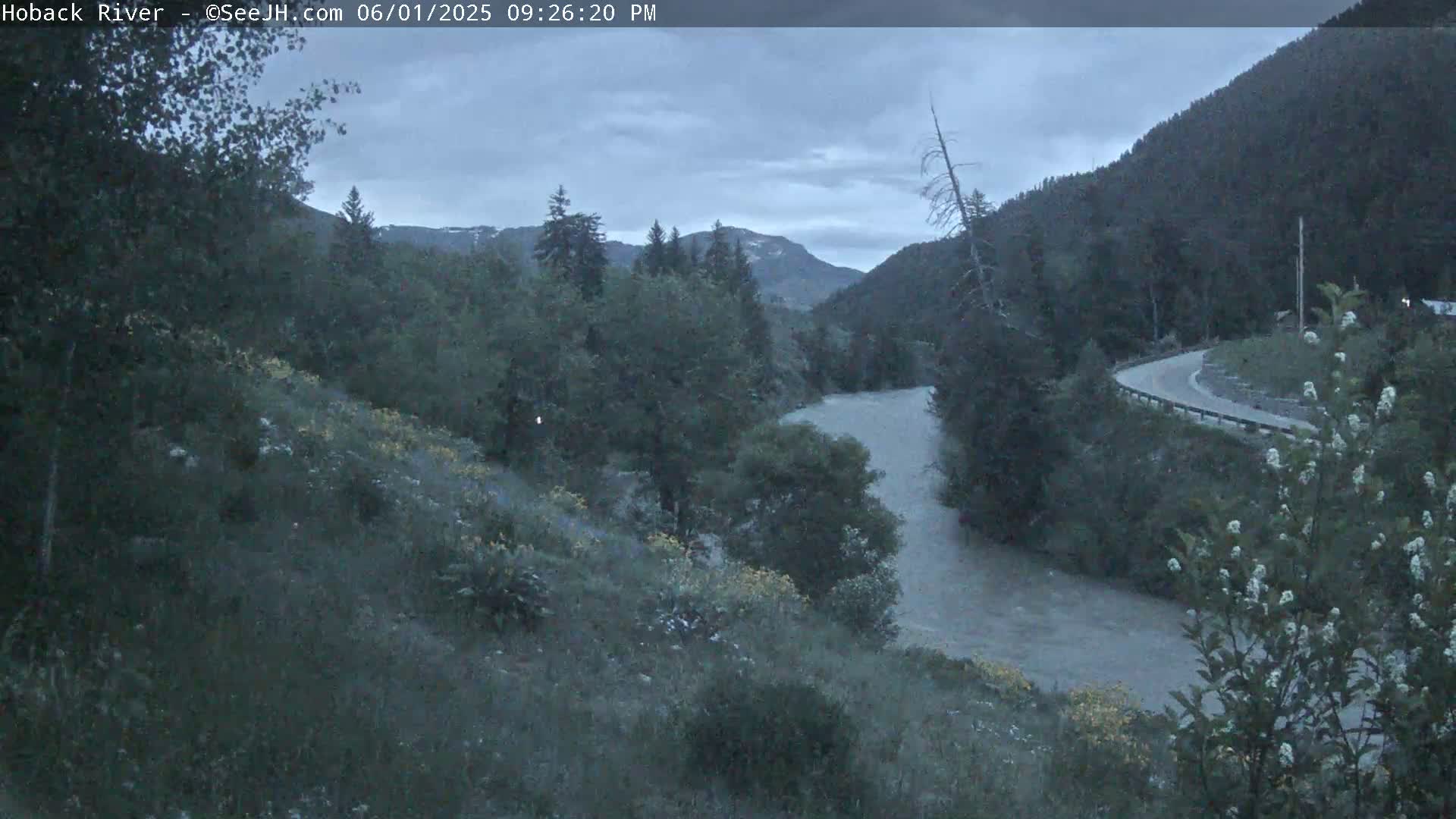 A river flows through a mountainous valley under a cloudy, dusky sky, with a road curving along the hillside in the background.