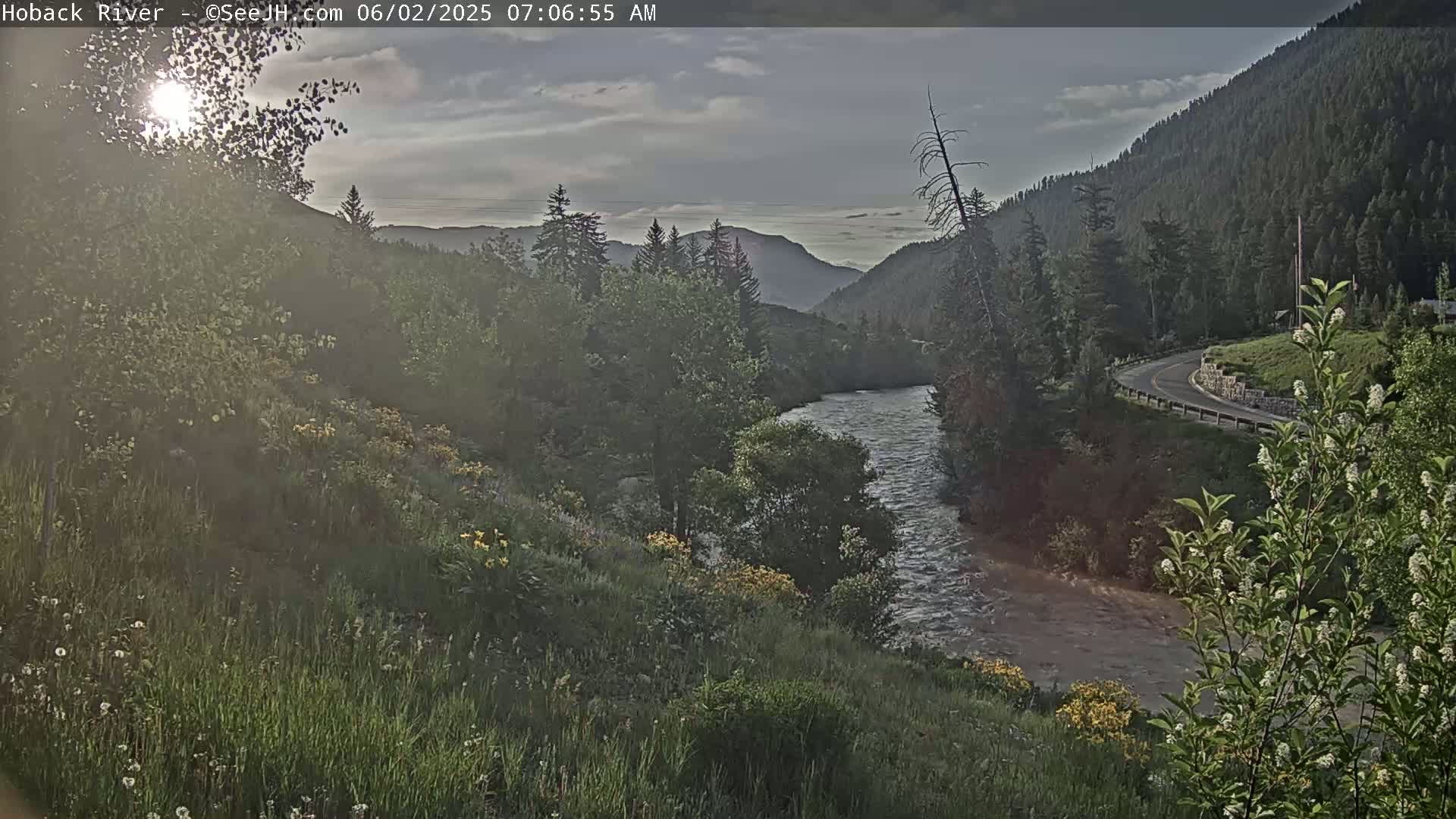 A sunlit river flows through a valley of green vegetation and trees under a partly cloudy sky, with a road curving along the mountainside in the background.