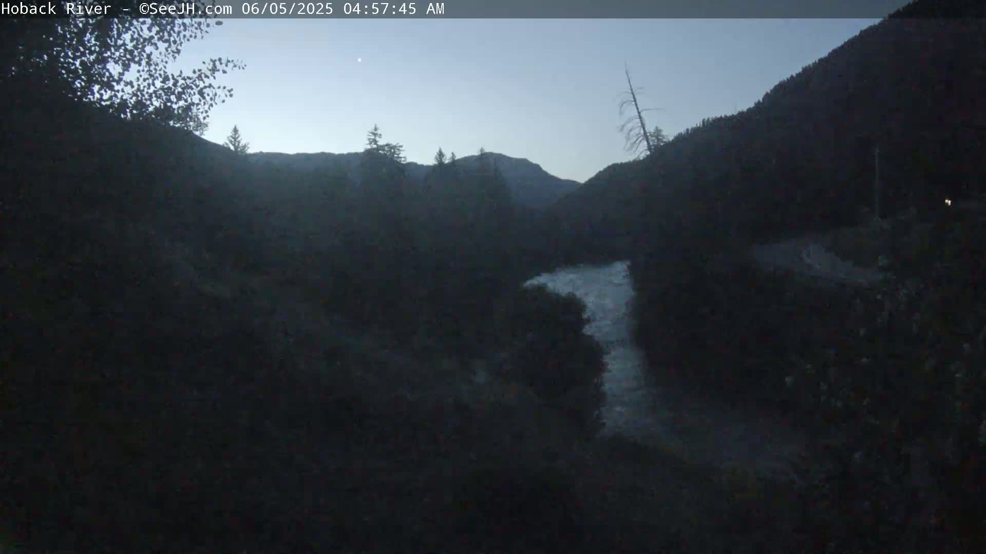 A dark, pre-dawn view shows a river winding through a mountain valley under a clear, twilight sky.