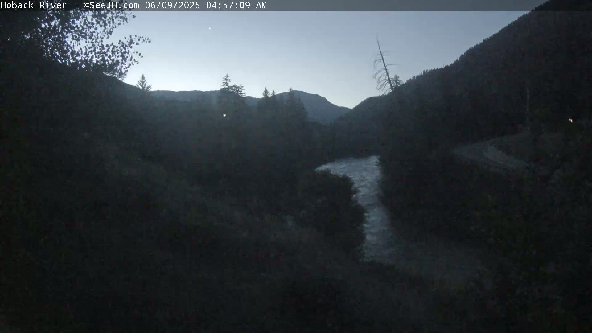 A dark, pre-dawn view shows a river winding through a valley surrounded by dark mountains and trees under a twilight sky.