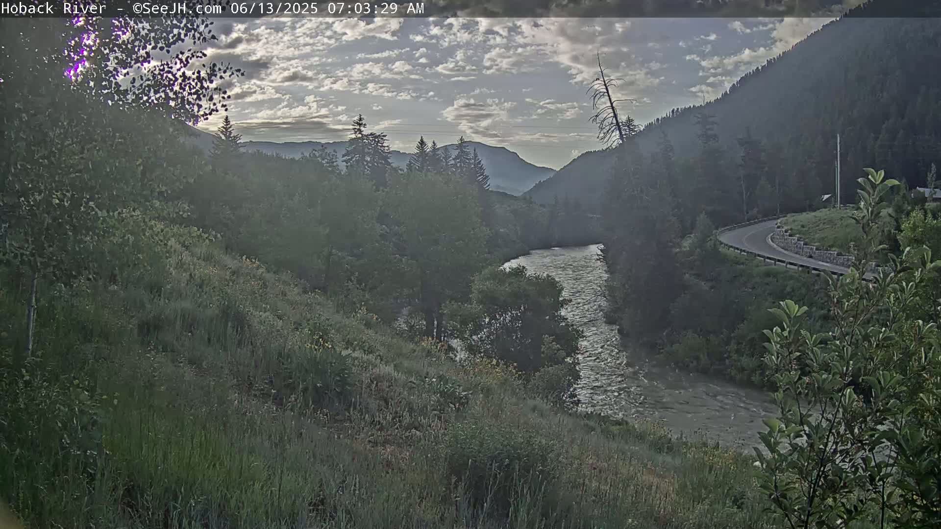 A river flows through a mountain valley under a partly cloudy sky at dawn.