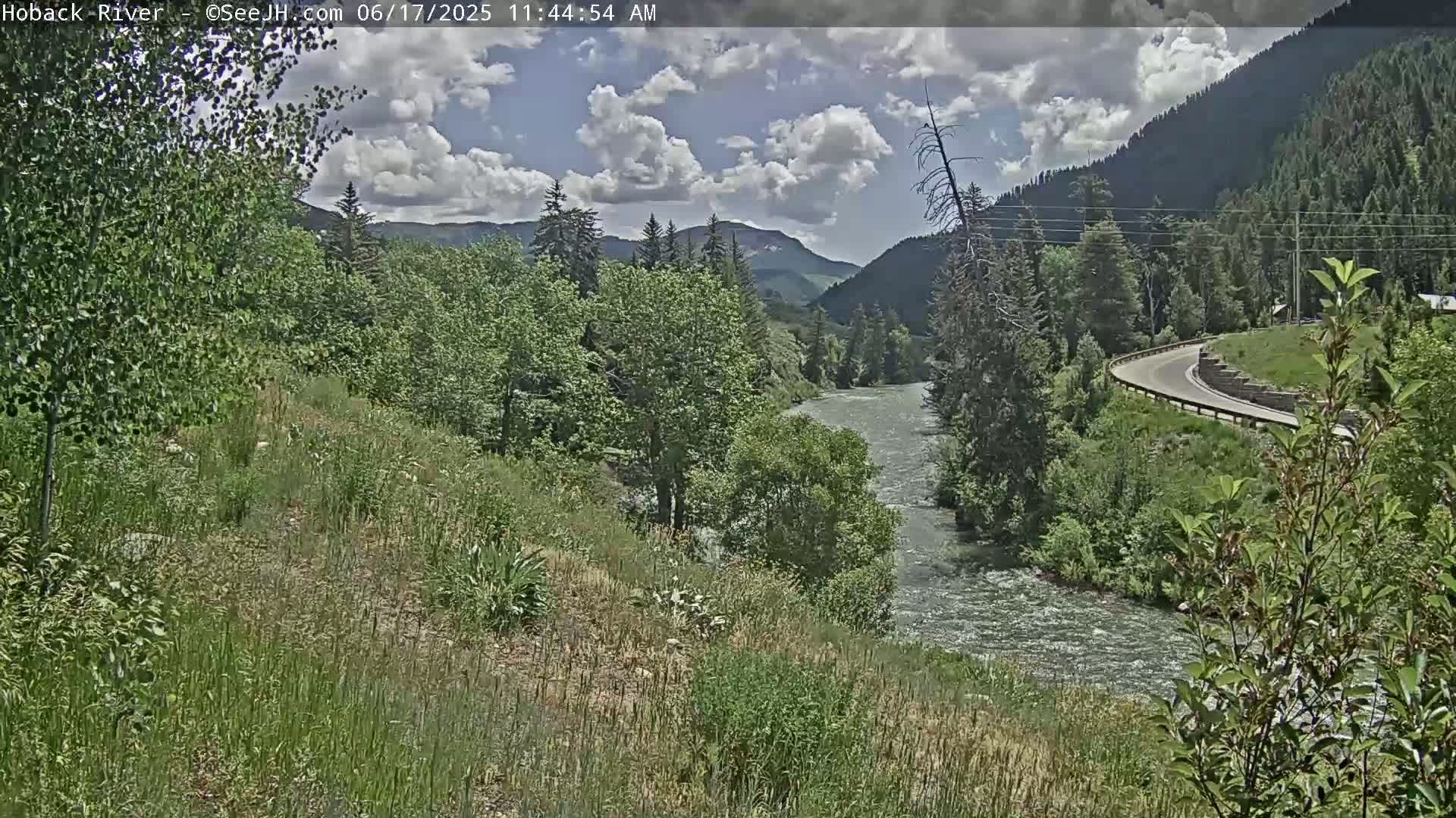 A partly cloudy day shows a flowing river winding through a lush green valley, bordered by mountains and a curved road.