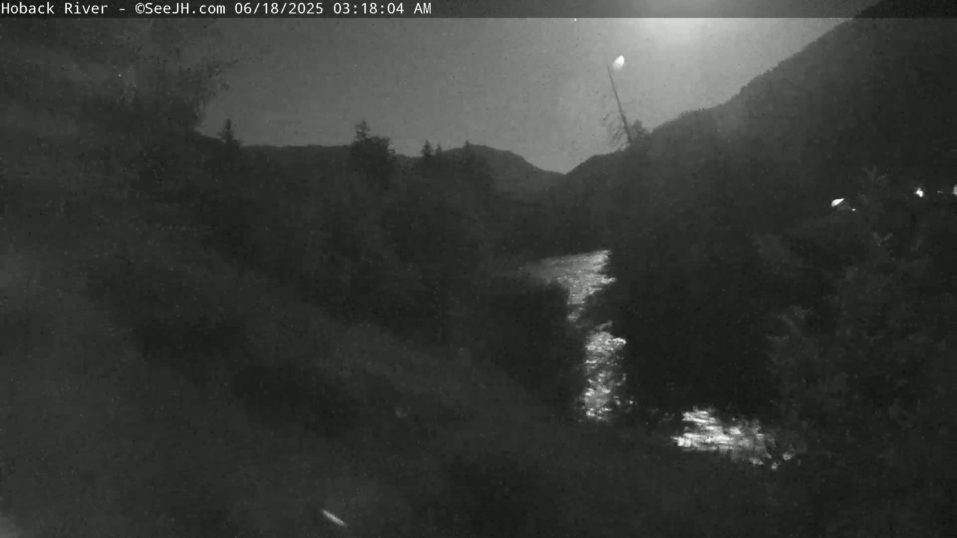 A nighttime, low-light view of a river flowing through a dark, mountainous landscape under a mostly clear sky.