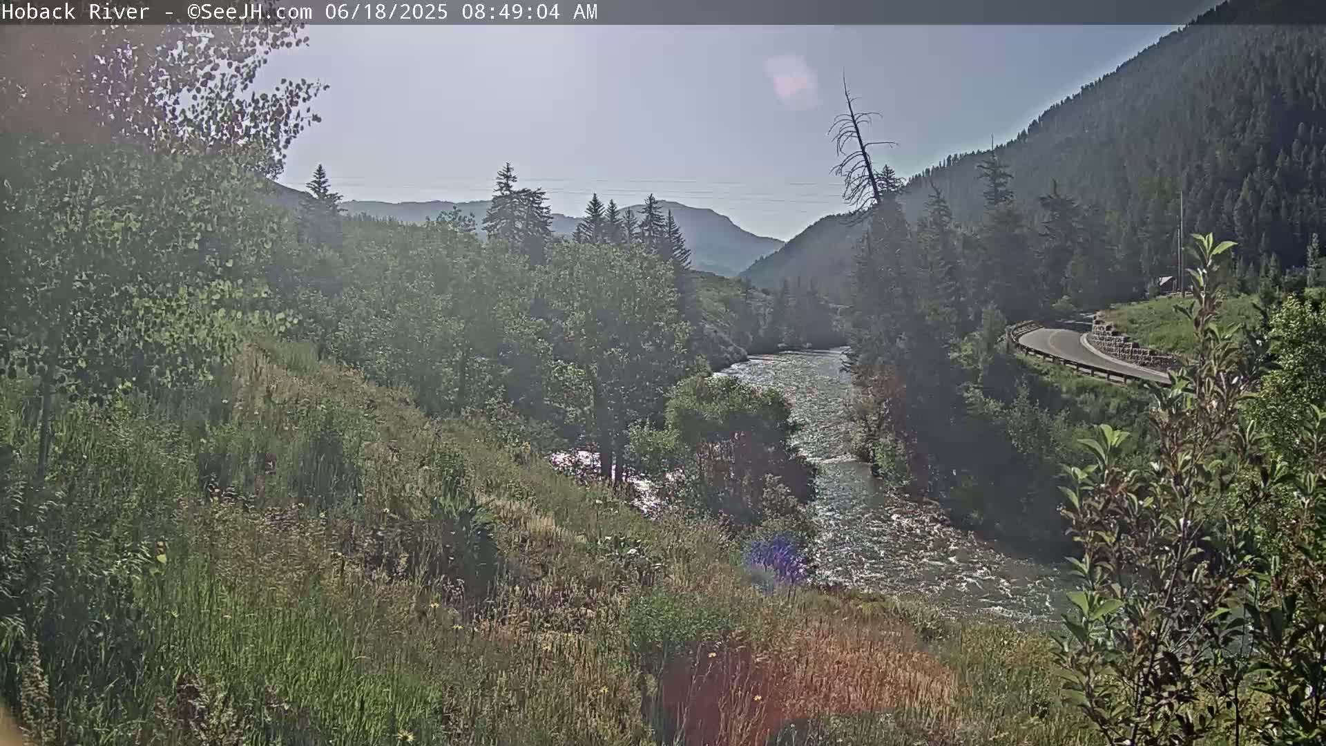 A river flows through a mountain valley on a hazy, sunny day, with a road curving along the mountainside.