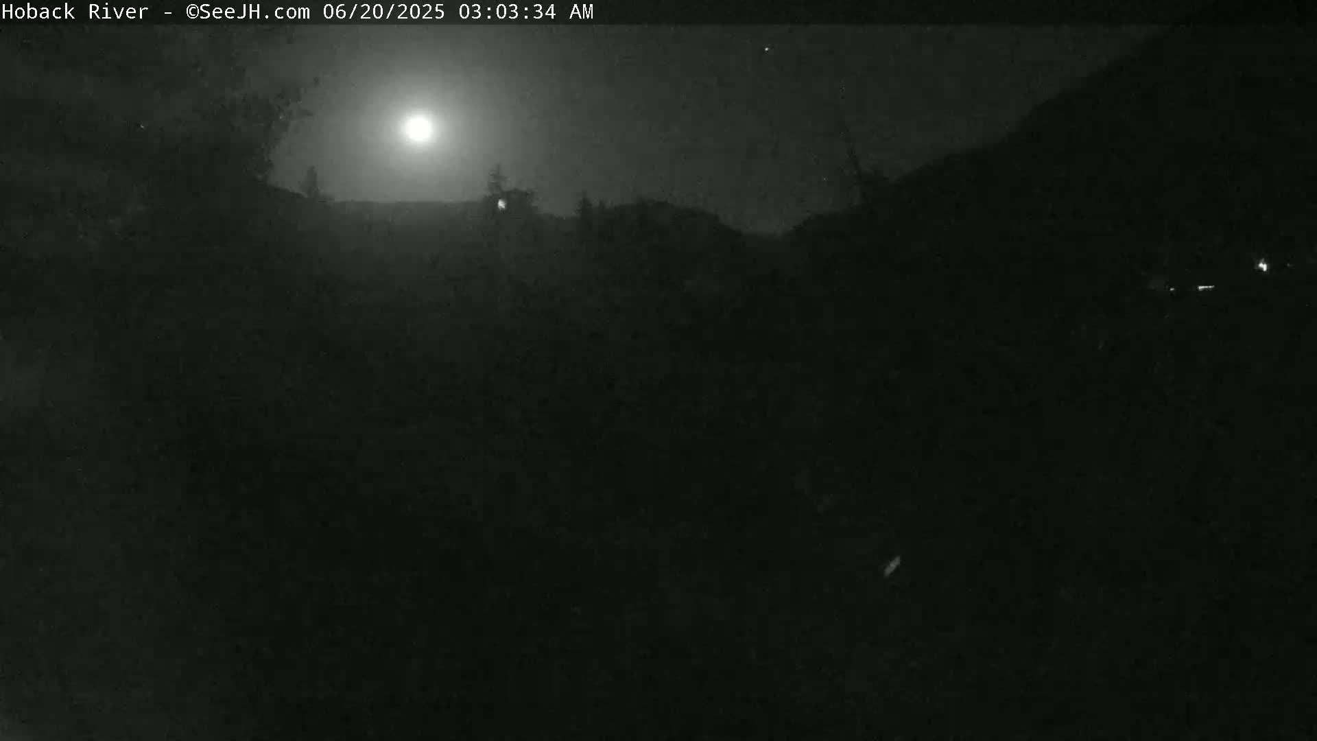 A nighttime, low-light outdoor scene shows a bright moon in the sky over dark, silhouetted trees and hills.