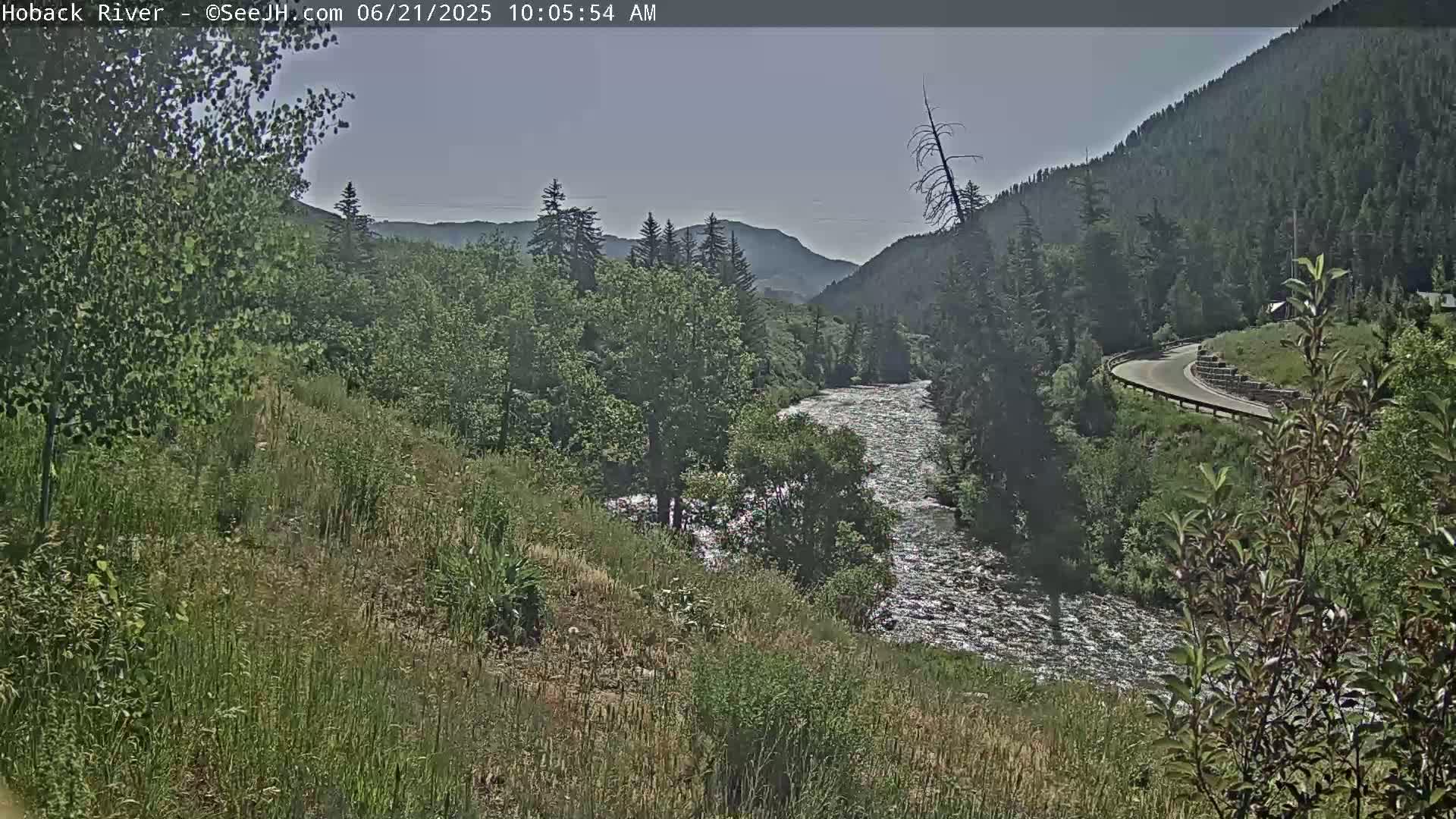 A fast-moving river flows through a mountainous valley on a partly sunny day.