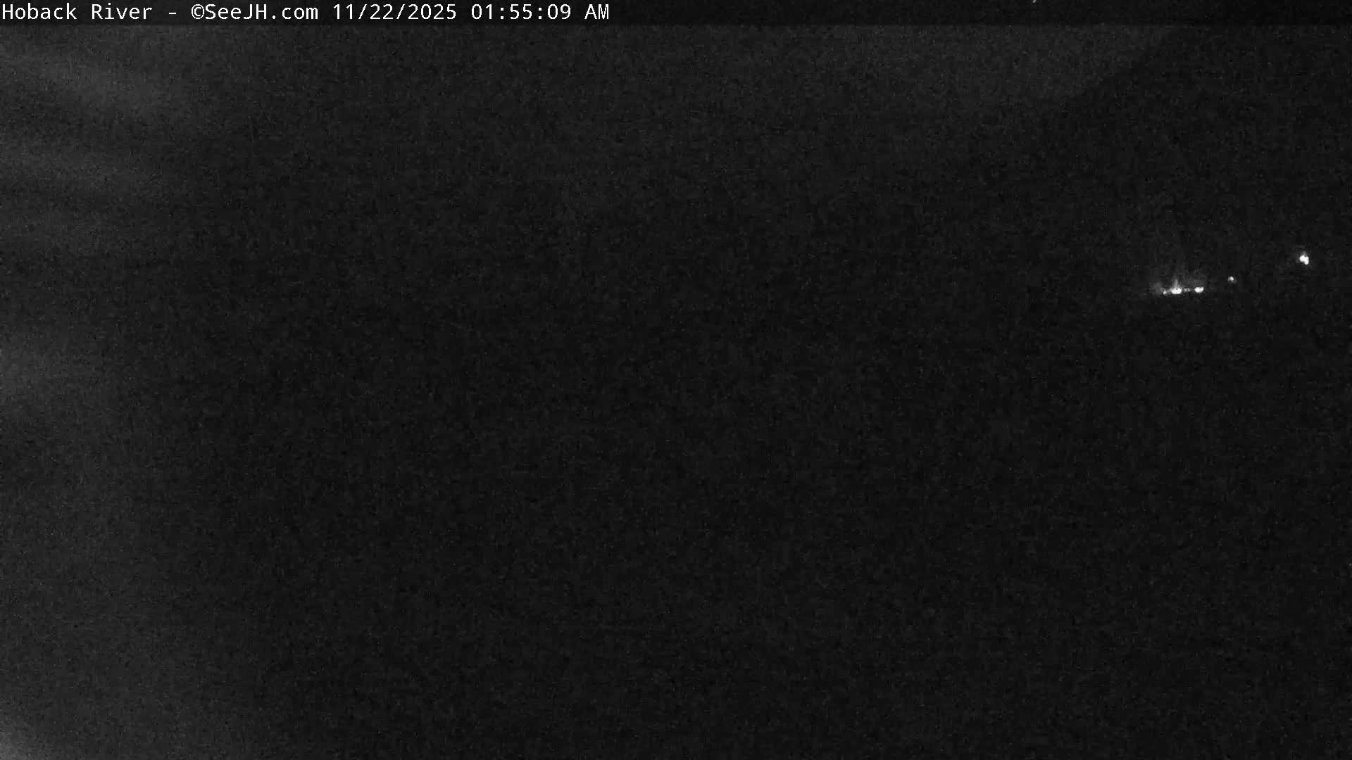 The image depicts an extremely dark outdoor scene at night with a few faint, scattered lights visible in the distance under clear skies.