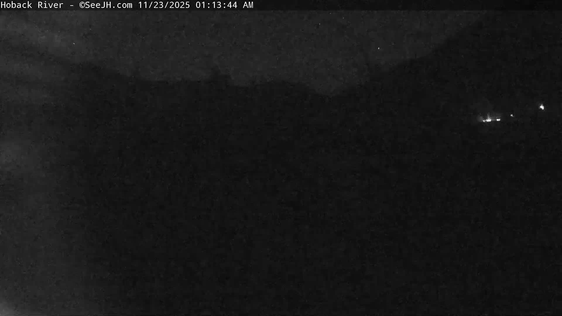 The image shows a very dark outdoor scene at night, with faint, indistinct land or cloud formations visible in the upper left and a cluster of small, bright lights in the upper right.