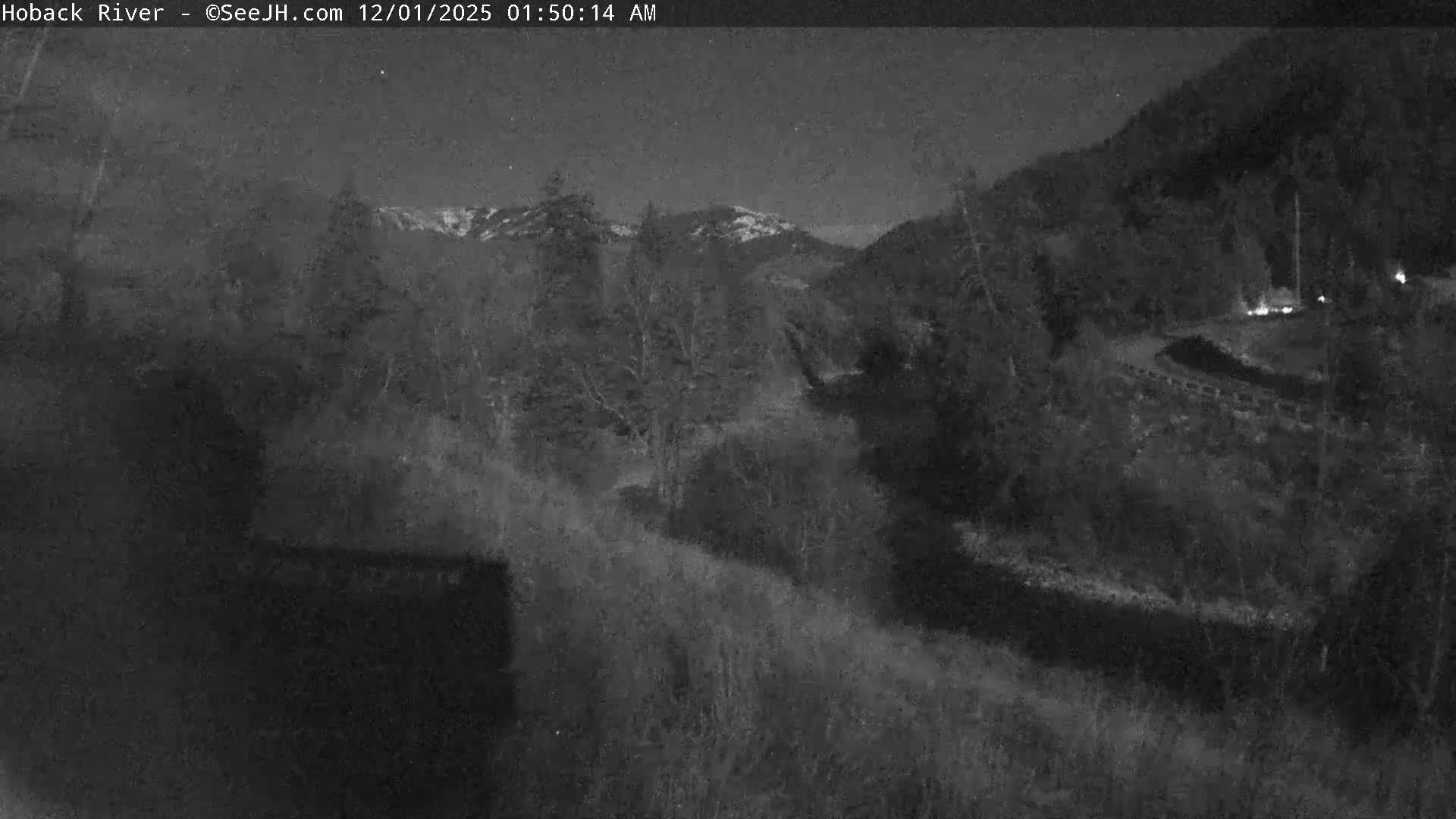 A clear, dark night reveals a river valley flanked by dense trees and distant snow-capped mountains under a starry sky, with a winding road and a few faint lights visible on the right.