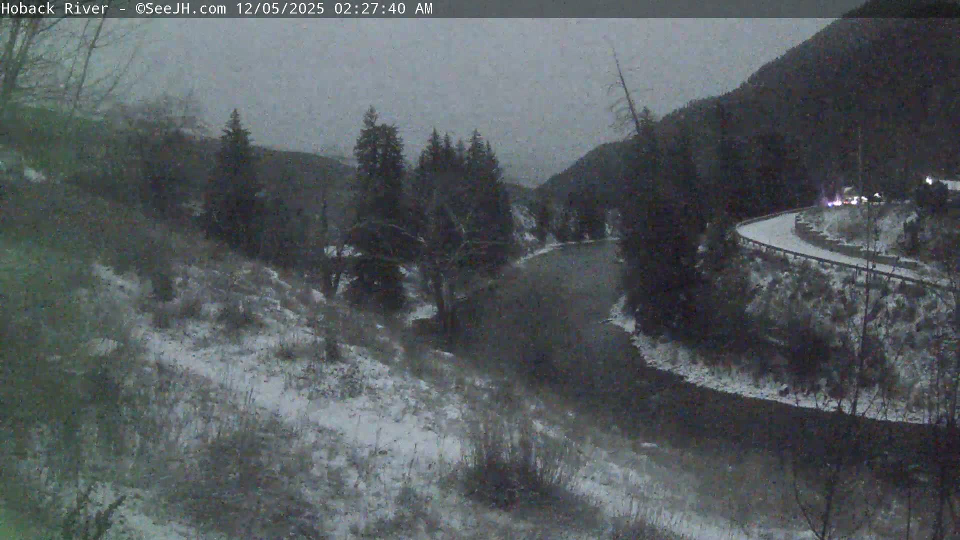 A dark, snowy winter landscape features a river meandering through snow-covered hills and evergreen trees, with a winding road and distant vehicle lights visible on the right.