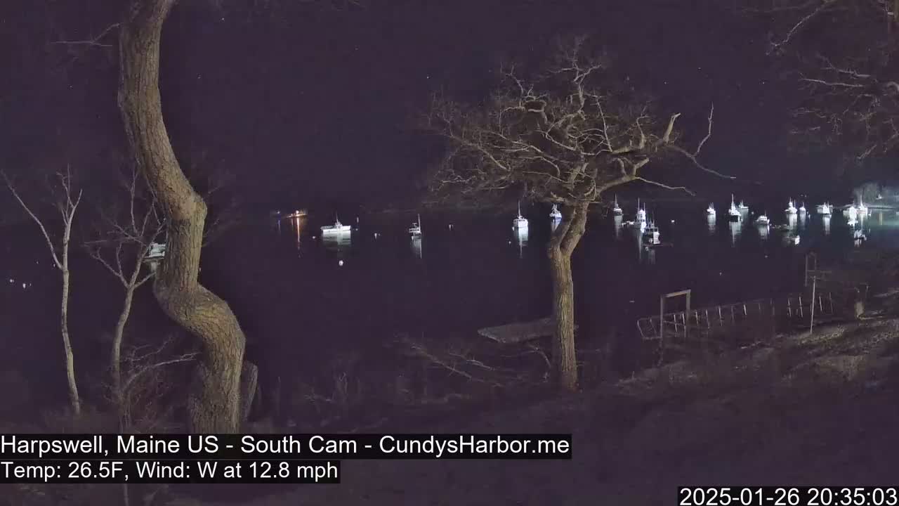 At night, under a dark sky with a 12.8 mph west wind and a temperature of 26.5°F, several boats are moored in a calm body of water, viewed from a shoreline with bare trees.