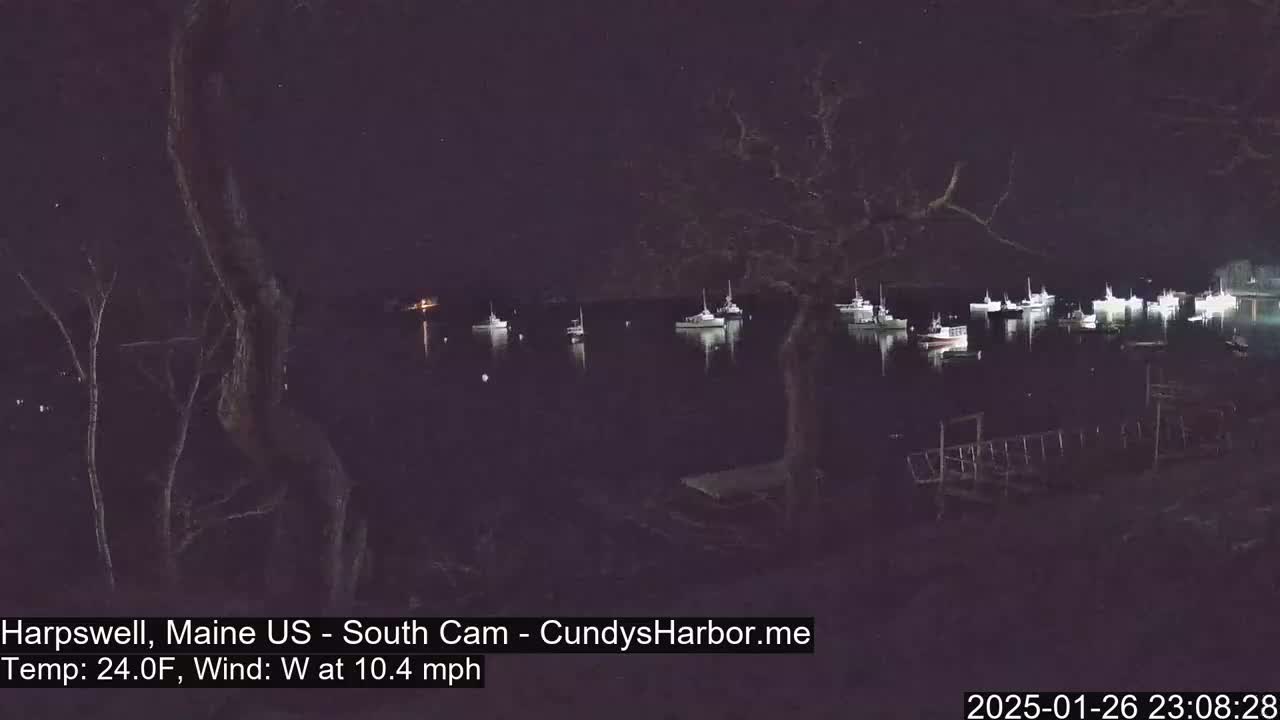 At night, under a clear sky with a westerly wind of 10.4 mph and a temperature of 24°F, several boats are moored in a calm harbor, observed from a dark shoreline.