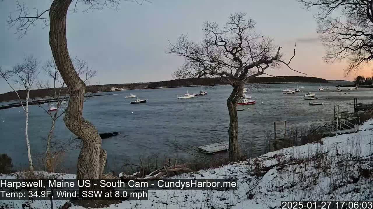 Several boats are moored in a calm bay under a twilight sky, with a light dusting of snow on the ground and bare trees in the foreground.