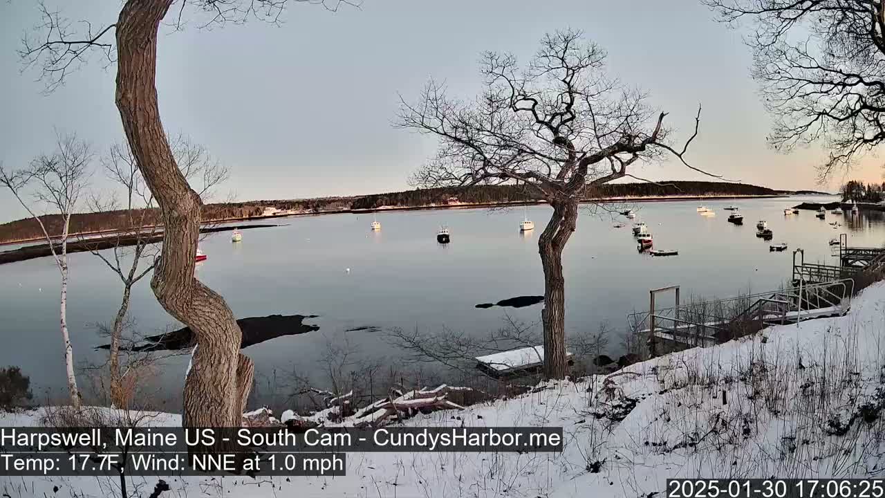 A snow-covered shoreline overlooks a calm bay with numerous boats, under a pale twilight sky.