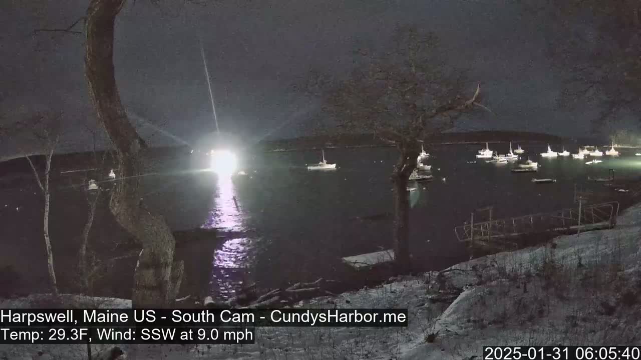 A nighttime view of a harbor with numerous boats, partially covered in snow, under a dark sky with a strong light source reflecting on the water.