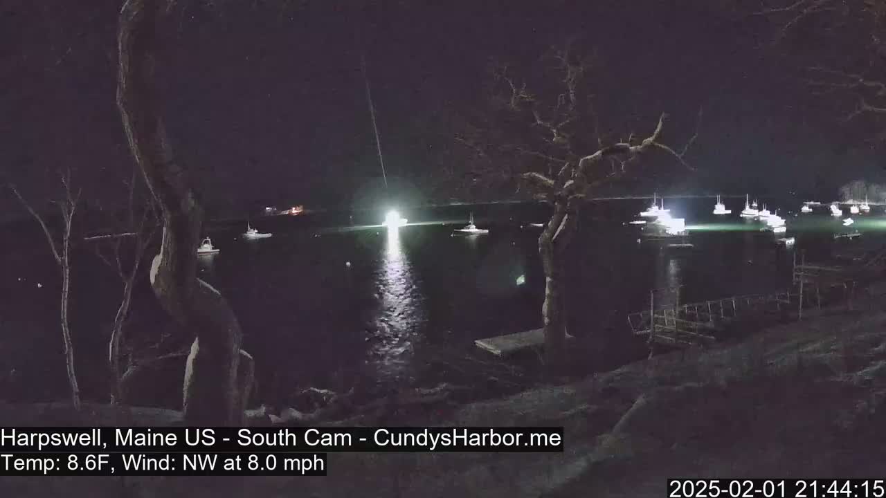 A nighttime view of a harbor with numerous boats illuminated, under a dark sky with a northwest wind at 8 mph and a temperature of 8.6°F.