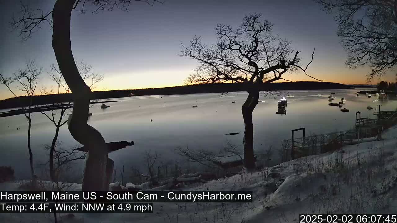 Several boats are moored in a calm, twilight harbor under a mostly clear sky, with snow on the ground and bare trees in the foreground.