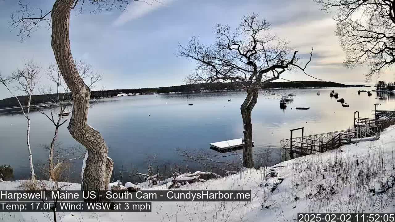 A snow-covered shoreline overlooks a calm bay with several boats, under a partly cloudy sky.