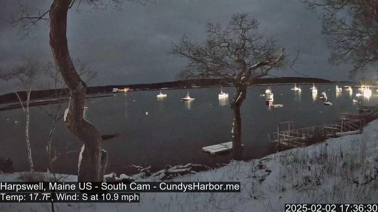 A snow-covered shoreline at dusk overlooks a calm harbor with numerous boats, under a dark, overcast sky.