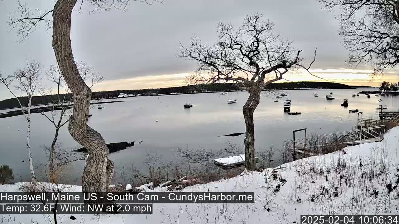 A snow-covered shoreline overlooks a calm bay with several small boats, under a mostly cloudy sky with a hint of sunset.
