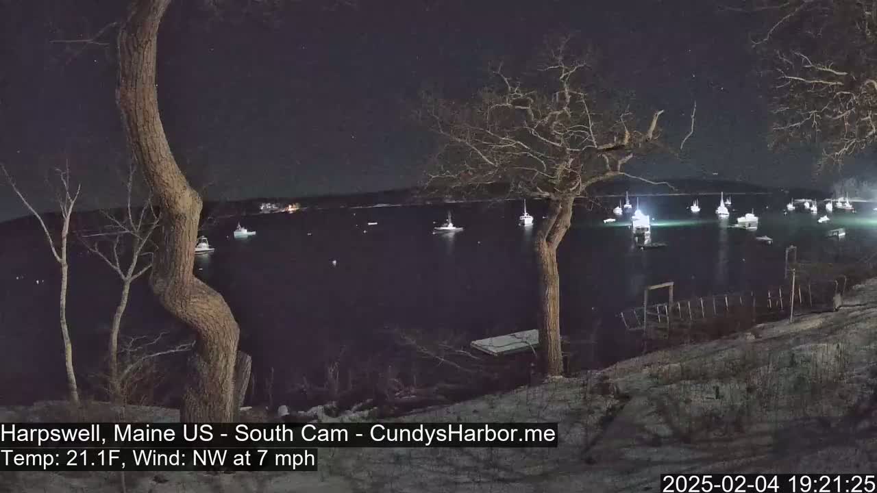 Several boats are moored in a dark harbor at night, under a clear sky, with a 7 mph northwest wind and a temperature of 21.1°F.