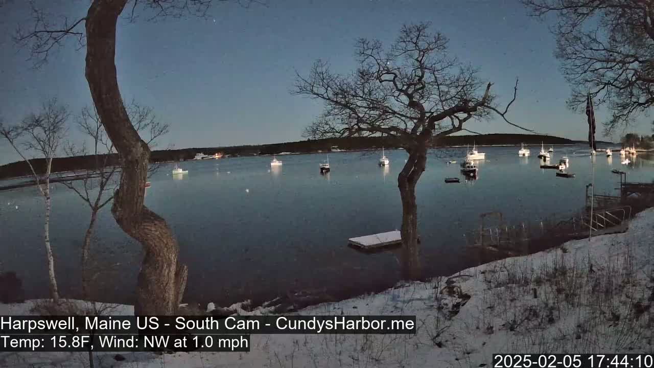 Several boats are moored in a calm harbor at twilight under a clear, dark sky, with a snow-covered shoreline and leafless trees in the foreground.