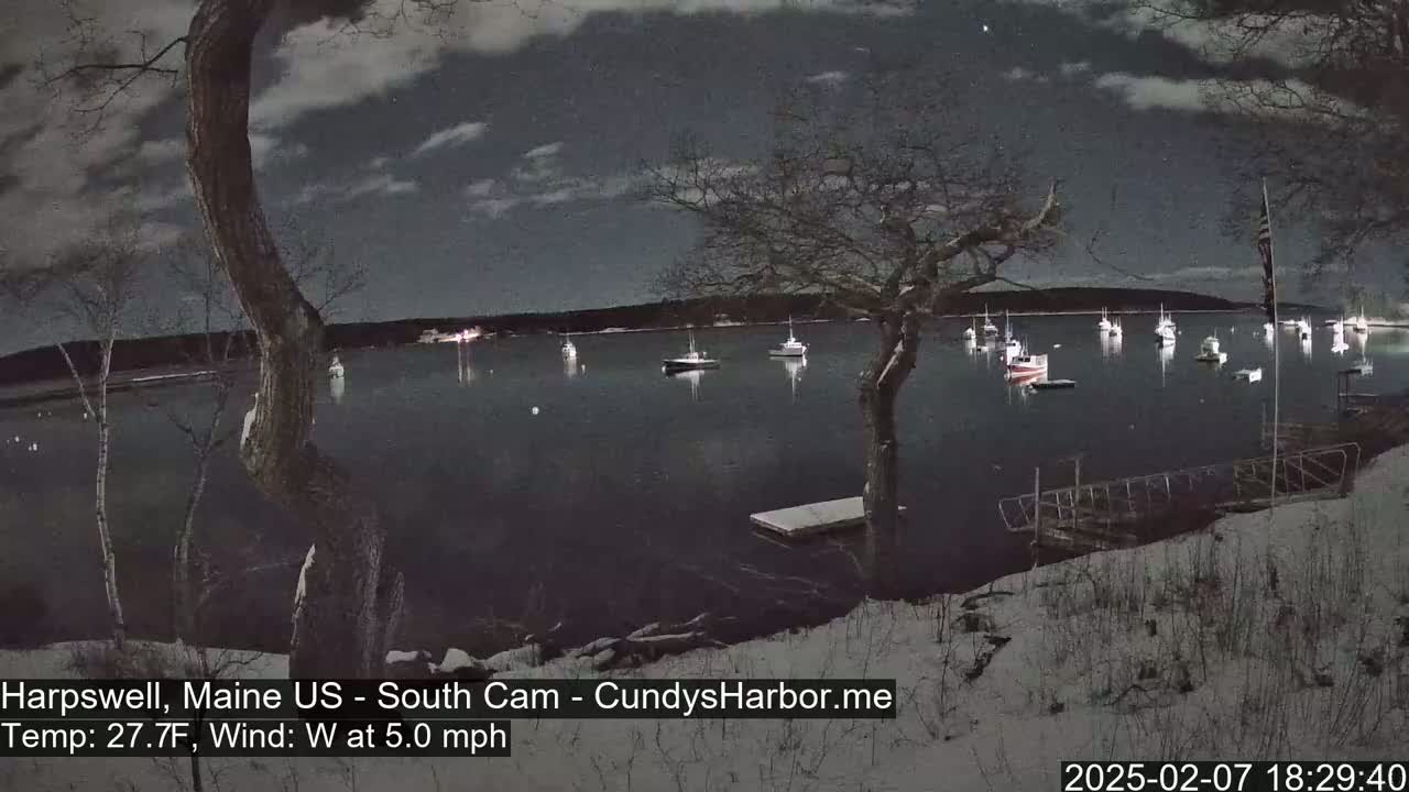 A snow-covered shoreline at night shows a calm body of water with numerous boats moored, under a partly cloudy sky with a light westerly wind.