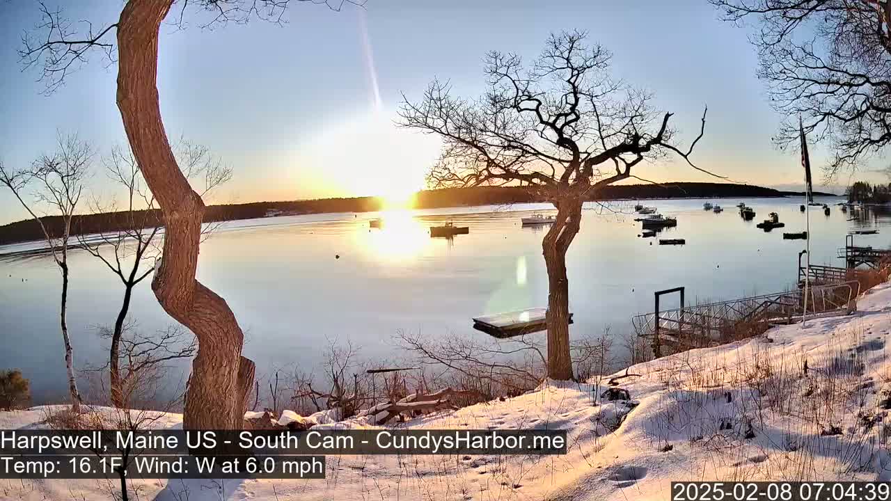 A snow-covered shoreline with leafless trees overlooks a calm bay at sunrise, where several boats are moored.