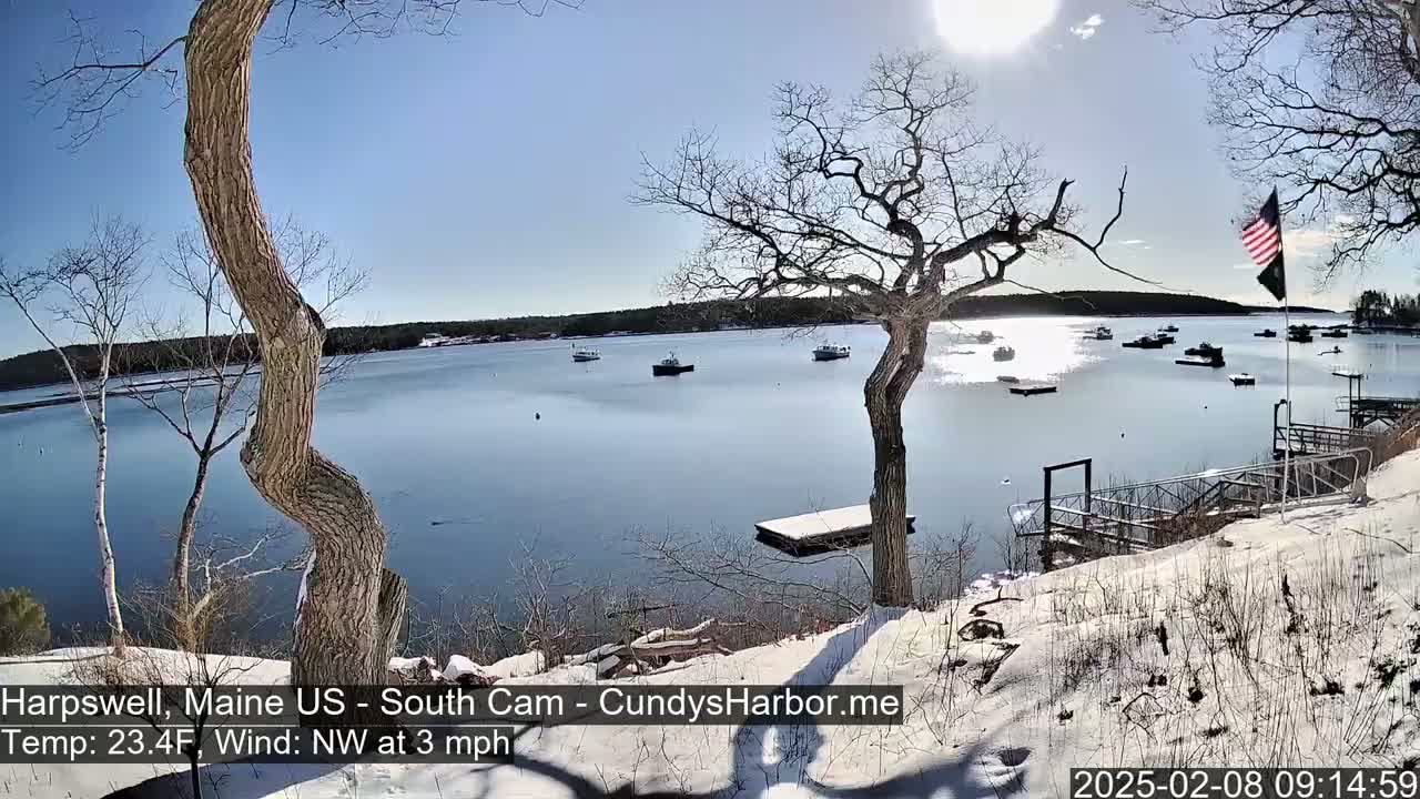A snow-covered shoreline overlooks a calm bay with several boats, under a sunny, partly cloudy sky.