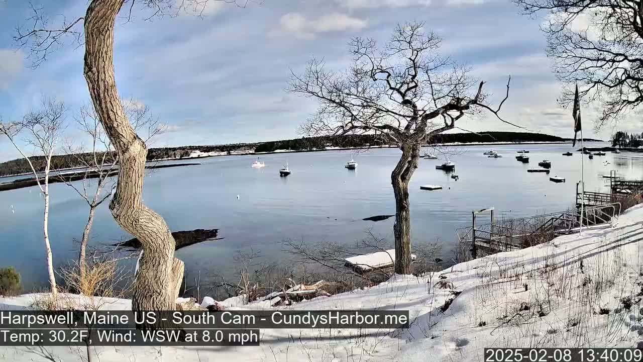 A snow-covered shoreline overlooks a calm bay with several boats moored under a partly cloudy sky.