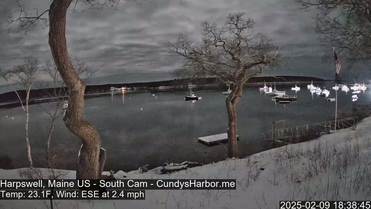 A snow-covered shoreline overlooks a calm, dark body of water where several boats are moored under a cloudy, gray night sky.