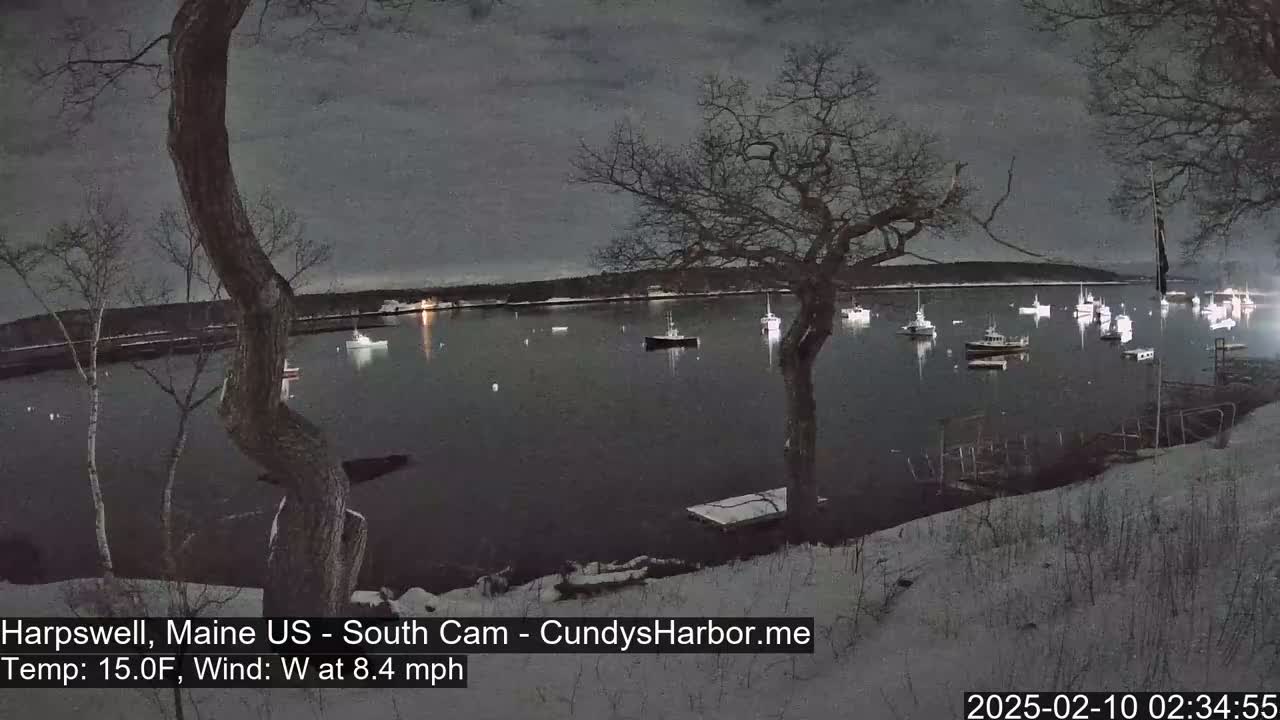 A snow-covered shoreline at night shows a calm harbor with numerous boats under a dark, overcast sky.