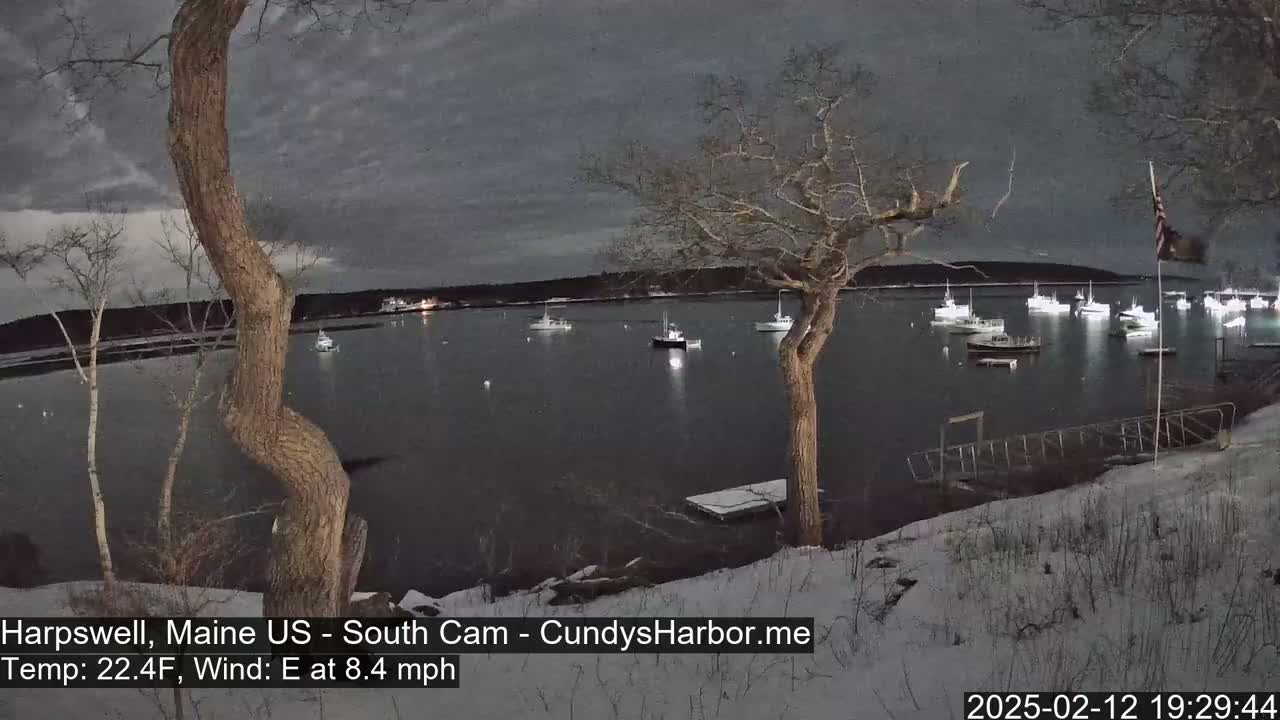 A snow-covered shoreline at night shows numerous boats in a harbor under a dark, cloudy sky with an eight-mile-per-hour easterly wind.