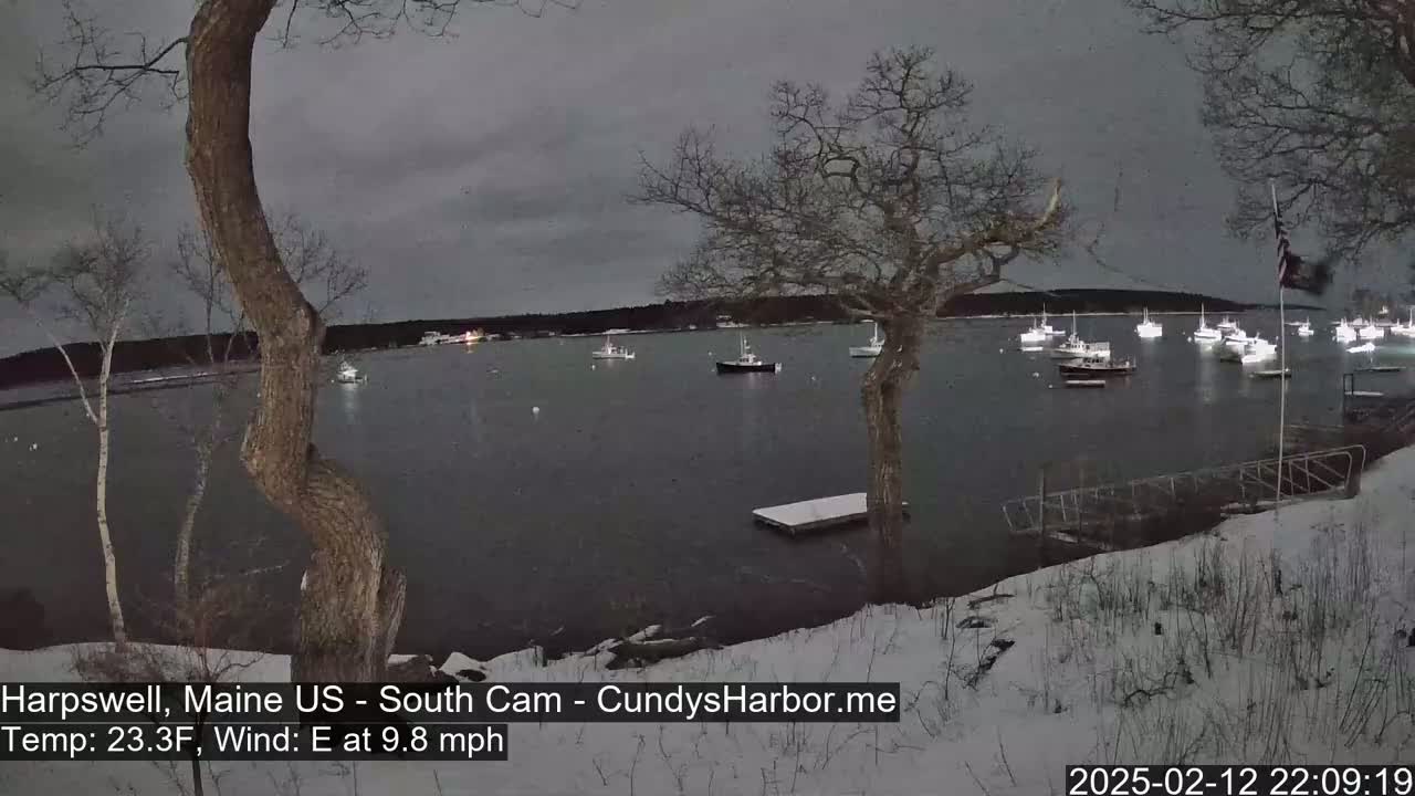 Several boats are moored in a harbor at night under a dark, overcast sky, with snow covering the ground in the foreground.