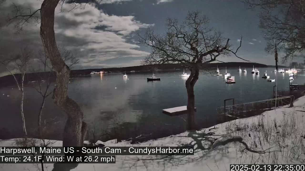 A moonlit harbor scene at night shows several boats moored in calm water under a partly cloudy sky, with snow on the ground and a 26.2 mph westerly wind.