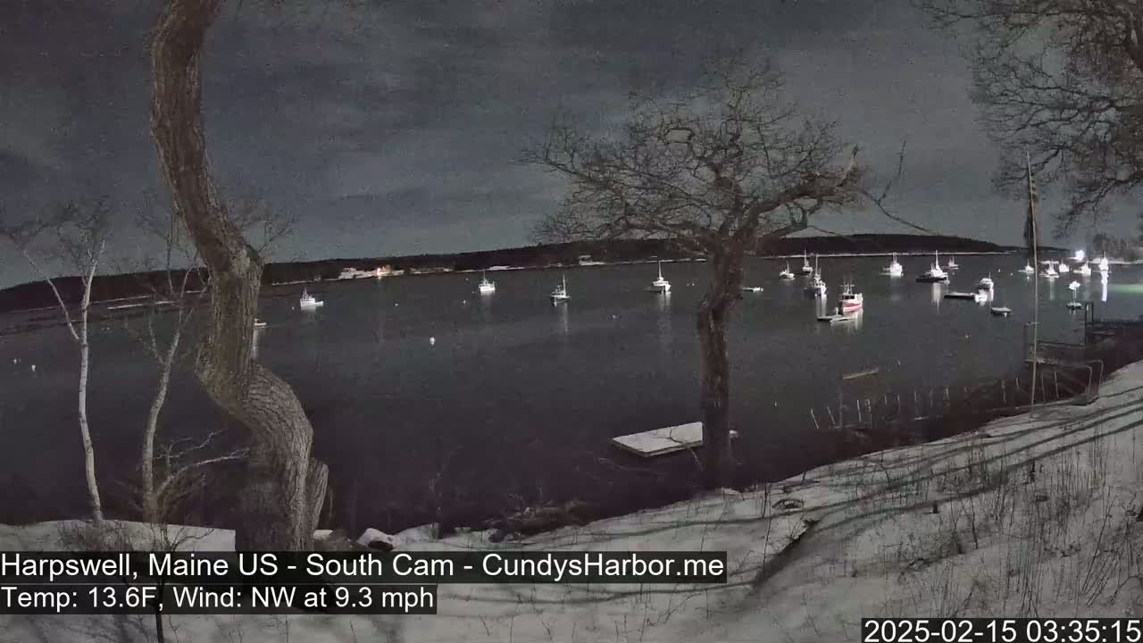 Under a dark, cloudy night sky, numerous boats are moored in a calm harbor, with a snowy shoreline in the foreground.