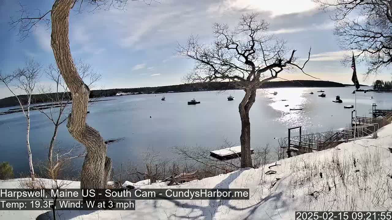 A snow-covered shoreline overlooks a calm bay with several small boats, under a partly sunny sky.