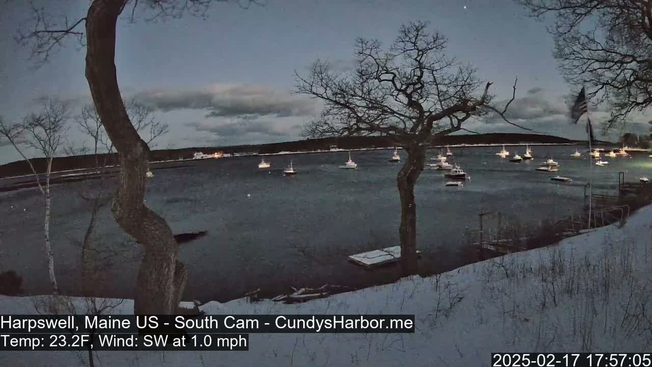 A snow-covered shoreline at dusk overlooks a calm body of water with numerous boats, under a partly cloudy sky.