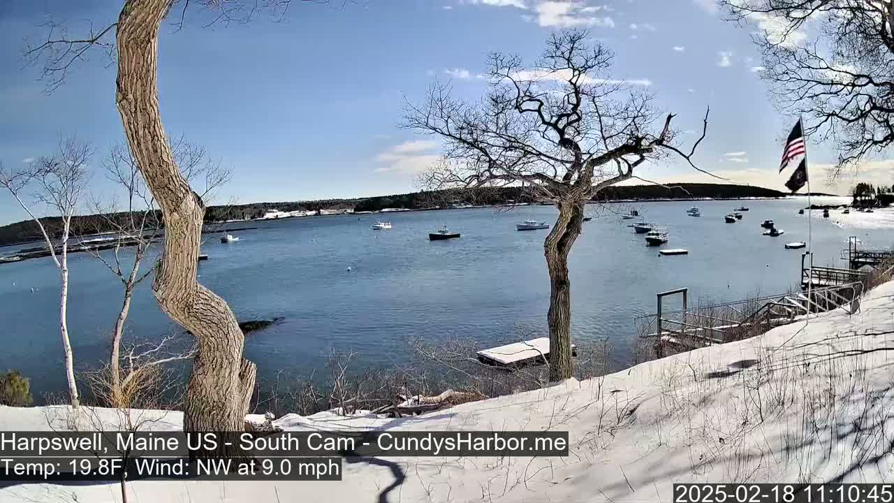 A snow-covered shoreline overlooks a calm bay with several boats under a partly sunny sky.