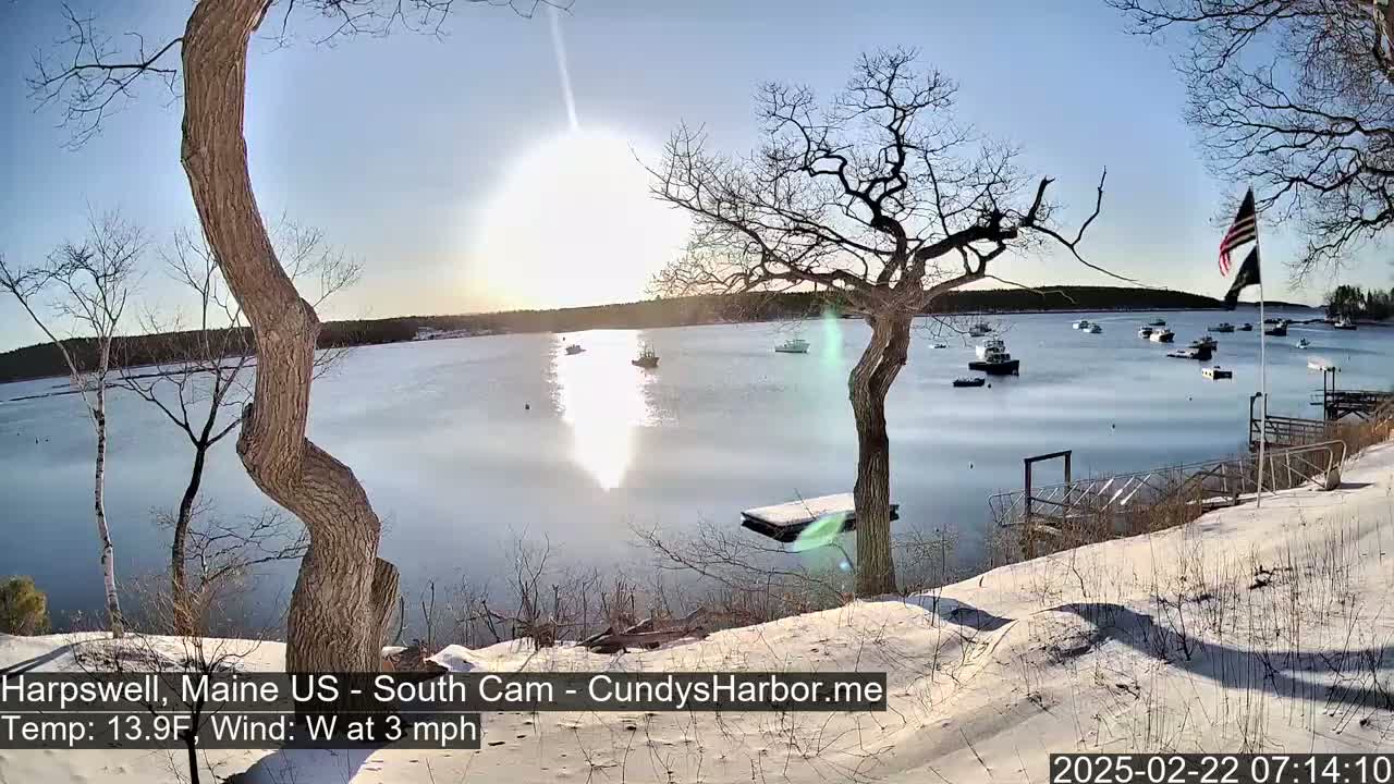 A snow-covered shoreline overlooks a calm bay with several boats, under a bright, sunny sky.