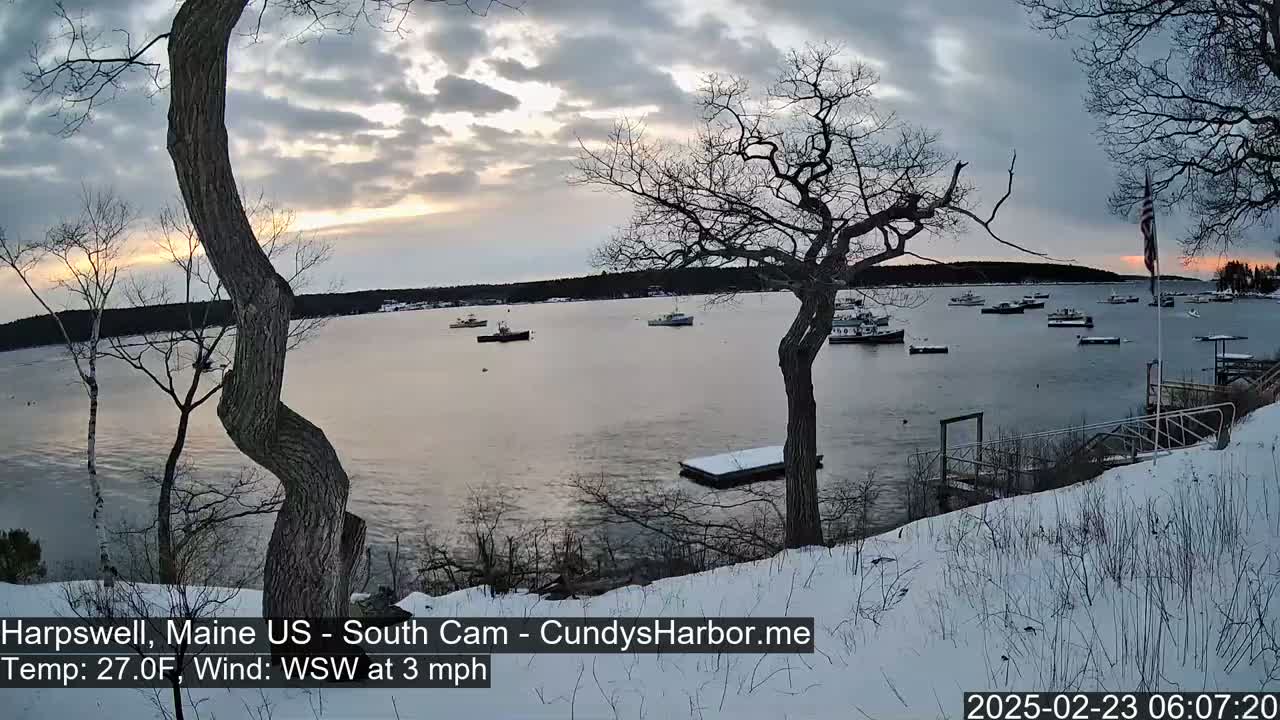 A snow-covered shoreline overlooks a calm harbor with several boats at sunset under a cloudy sky.