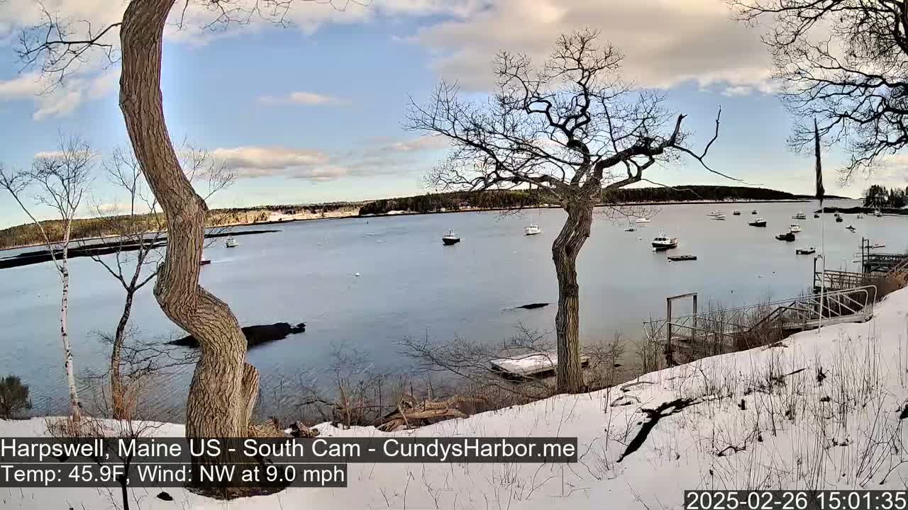 A calm bay with several small boats is seen from a snow-covered shoreline, under a partly cloudy sky.