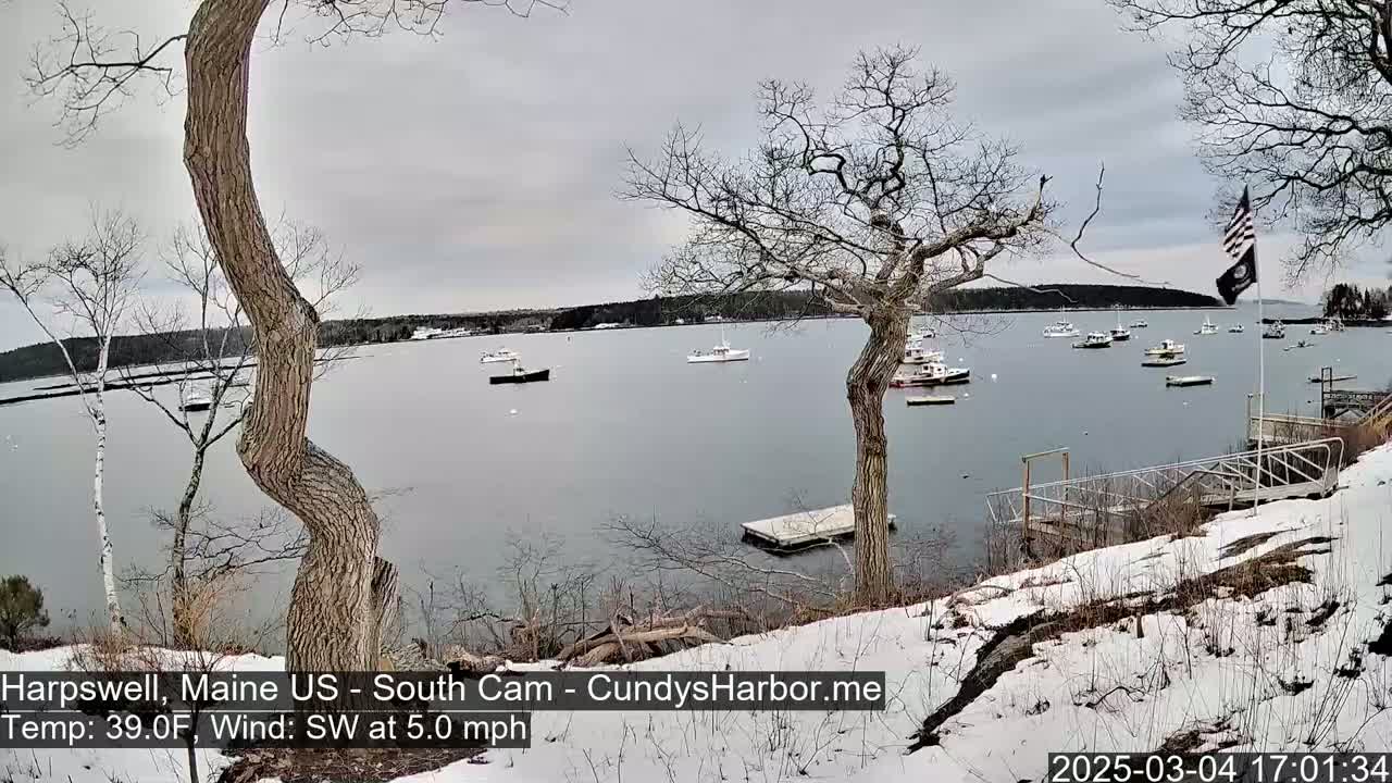 A snow-dusted shoreline with several boats in a calm bay under a cloudy sky.