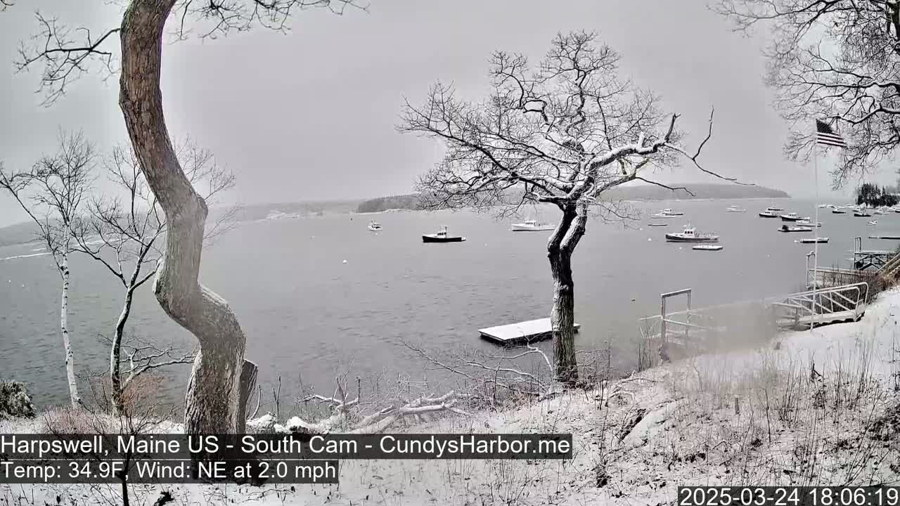 A snow-covered shoreline with several boats in a calm bay under a gray sky.